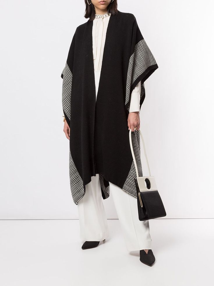 a woman is standing in front of a white background. she is wearing a black and white striped poncho and white pants. she is carrying a black and white handbag with silver accents. she is wearing black shoes.