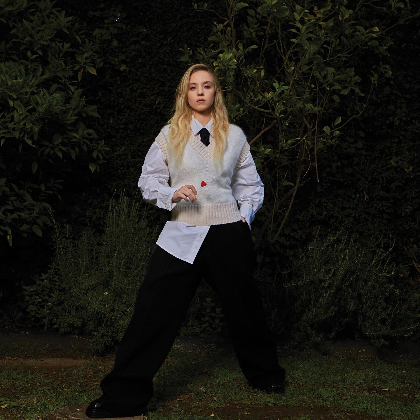 a fullbody shot of a young blonde woman posing outdoors in a garden setting.  she's dressed in a stylish outfit consisting of a creamcolored knitted vest, a white oversized buttondown shirt, and black wideleg trousers. she wears a black tie under her shirt and has a serious, neutral expression on her face. the background is dark and lush, consisting of dense green bushes and trees, creating a contrast with her lightcolored outfit. the overall aesthetic is elegant and chic.