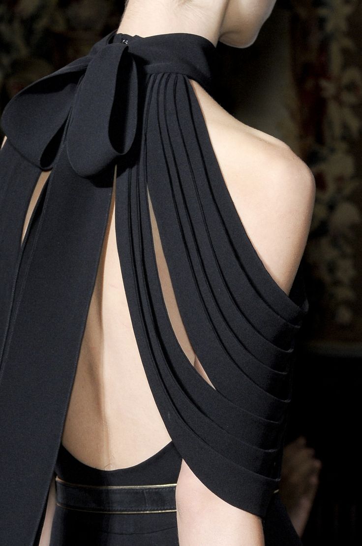loseup view of a woman's back, showcasing a striking black dress. the dress features a high halter neck with a large bow detail at the nape of the neck.  the back is completely open, revealing the model's back. the fabric of the dress is pleated and draped, creating a sophisticated and elegant look. the dress appears to be made of a structured fabric, perhaps silk or crepe.  the background is blurred but suggests a highfashion setting, possibly a runway or photoshoot. the overall impression is one of luxury and highend fashion.