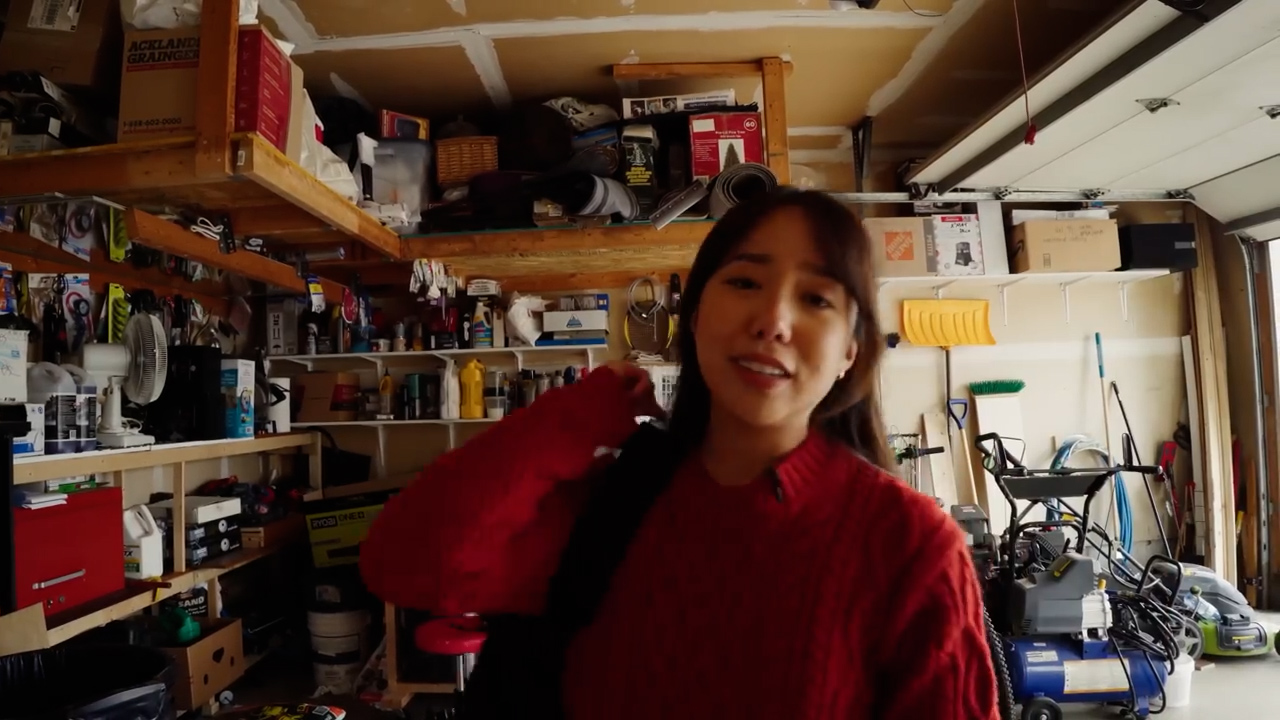 a young woman stands inside a cluttered garage while talking to the camera. she is wearing a red sweater and has a backpack slung over her shoulder. the garage is filled with shelves and various items.