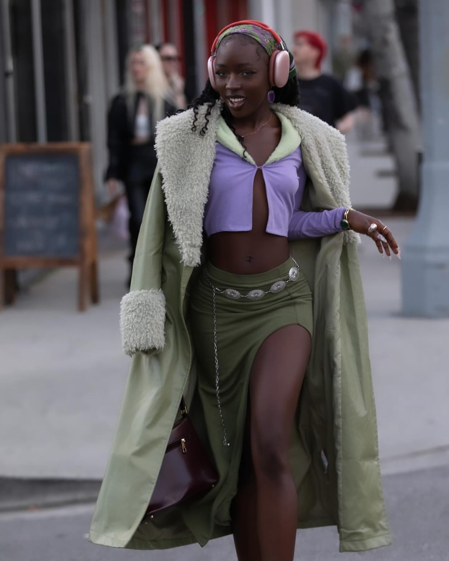 a woman in a green coat, a purple crop top and a green skirt with a slit walks down the street. she wears a silver chain belt, a brown bag and headphones.  her hair is styled in a curly look and she is wearing makeup.