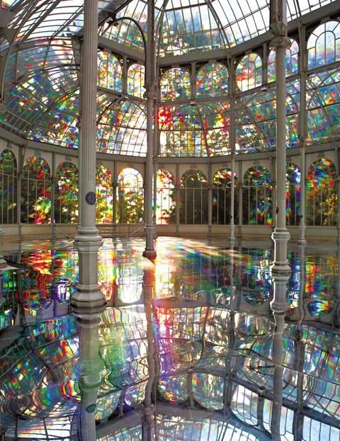 the image showcases the interior of a glassenclosed structure, possibly the crystal palace in madrid, spain.  the entire space is filled with an installation of vibrant, rainbowhued stained glass, both in the windows and potentially incorporated into the floor as well. the floor acts as a massive mirror, reflecting the stunning colorful light and architectural features of the building. sunlight streams in, creating dazzling reflections and intensifying the kaleidoscopic effect.  the architectural details, particularly the white columns and intricate framework, are visible through the colorful glass and their reflections. the overall impression is one of vibrant beauty and artistic sophistication.