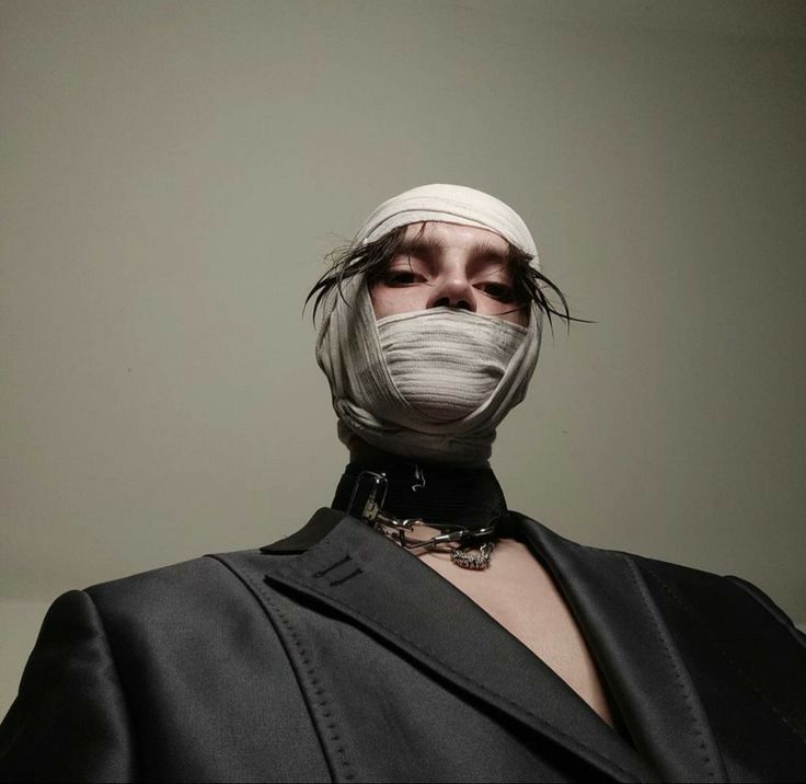 a young man with a bandaged face, a black choker, and a black suit is looking intensely at the camera.  he is wearing a bandage over his face that covers his entire head except for his eyes and eyebrows. he has short black hair and a serious expression on his face. he is wearing a black blazer and a black choker with a silver necklace. he has an intense stare and an edgy style that is both elegant and dramatic.