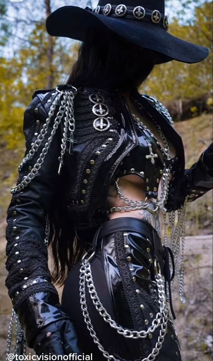 a woman in a black leather jacket and black pants with lots of chains and metal accessories. she is wearing a wide brimmed hat with an upside down pentagram design around the brim. the woman is looking off to the side.
