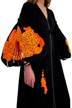 a woman is wearing a black longsleeved dress with orange floral embroidery. the dress has a vneckline and a belt. the sleeves are puffy and have the floral design on them. she is standing in a studio with a white background.