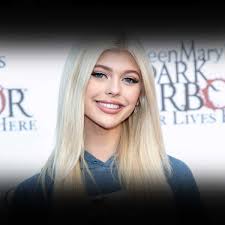 a closeup shot of loren gray smiling at an event. she has blonde hair and is wearing a blue top. the backdrop behind her features event signage with lettering in white and red.