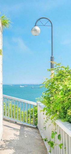 this is an idyllic outdoor scene overlooking the ocean on a bright sunny day. the image features lush green plants, a white railing, and a lamp post with a round globe. the blue sky and sea evoke a sense of tranquility and vacation vibes.