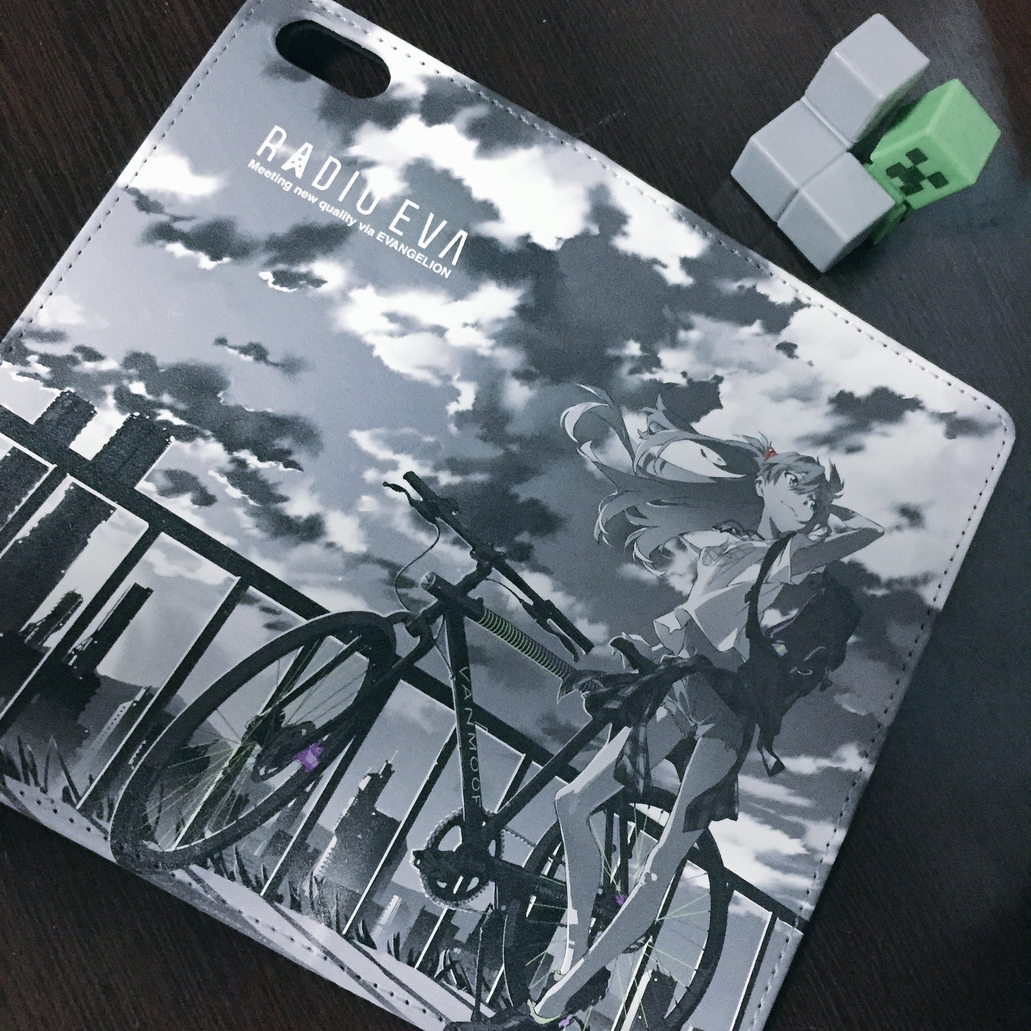 a black and white radio eva phone case with an image of asuka langley soryu from neon genesis evangelion leaning on a bike. the background features a cityscape. two minecraft foam bricks are also visible in the top right corner.