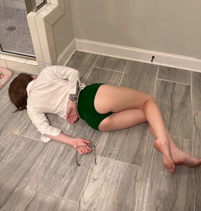 a woman wearing a white shirt and green shorts is lying on a tiled floor in a bathroom. her glasses are next to her hand.