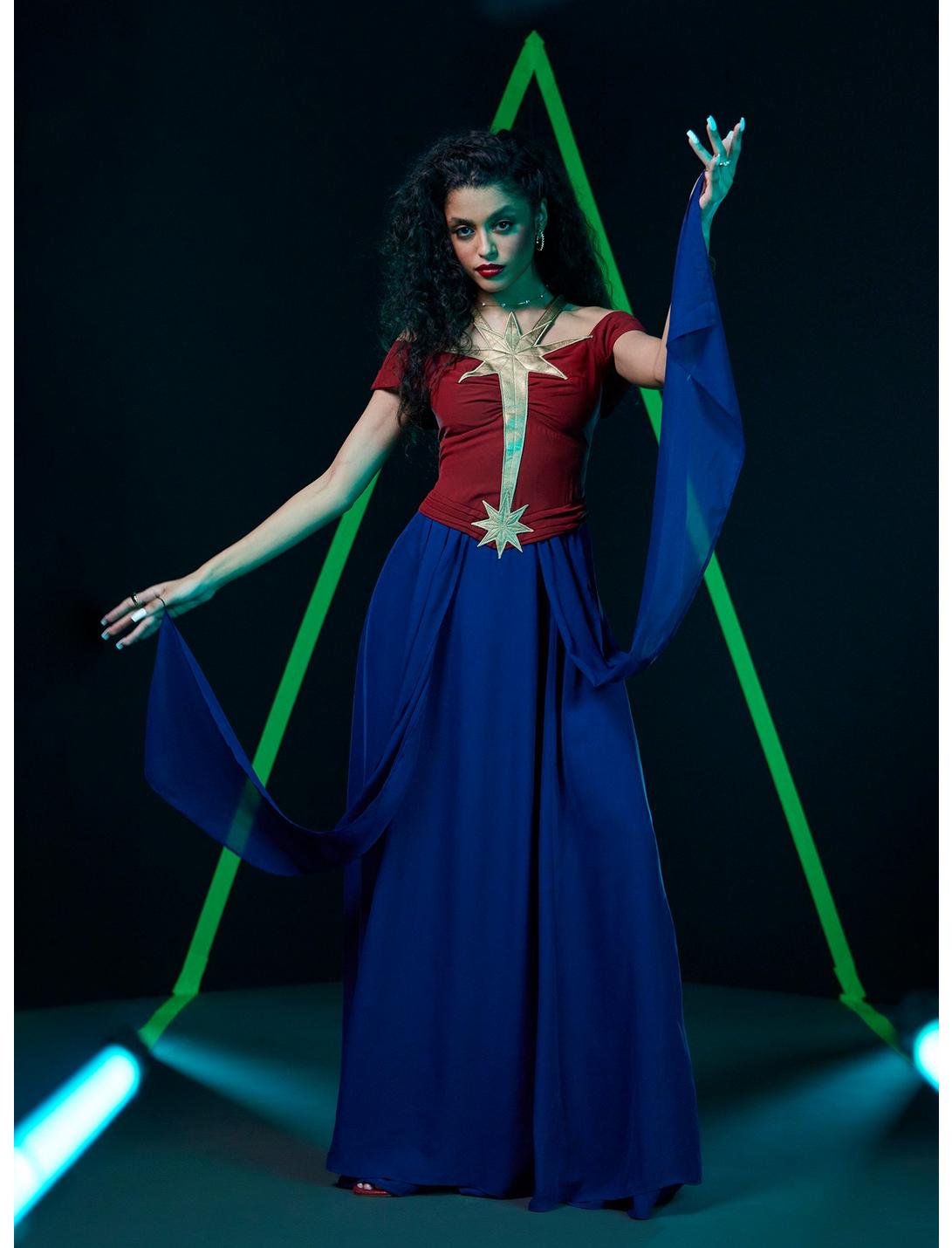 a woman is dressed in a theatrical costume. she is wearing a maroon top with gold accents, a floorlength dark blue dress, and flowing blue fabric draped from her arms. she has dark curly hair, red lipstick, and is wearing jewelry. the background is a dark stage with a neon green triangle and stage lighting. she is striking a pose and looking towards the camera.