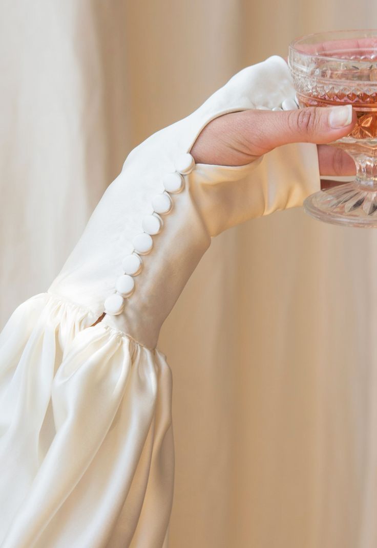 an upclose shot depicts the hand of a person holding a glass of champagne while wearing a creamy white dress with a sleeve lined with scalloped buttons.