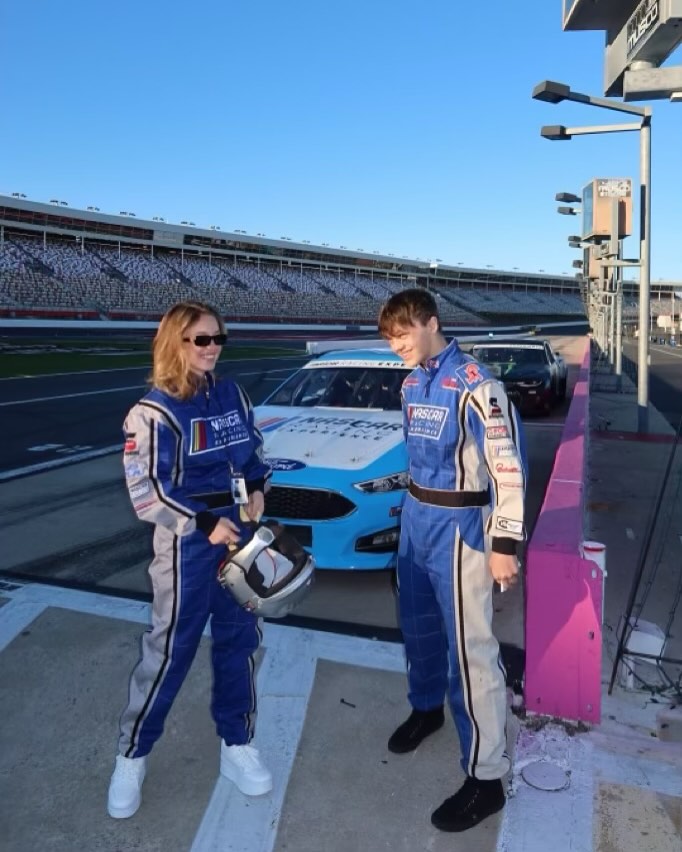 the image shows a young man and woman, likely teenagers, dressed in nascar racing suits standing next to a blue ford race car at what appears to be a nascar race track. the background features a large stadium and pit lane. the woman is holding a racing helmet.  the overall mood suggests a behindthescenes or prerace moment at a nascar event. the sun is shining brightly, indicating a daytime setting.