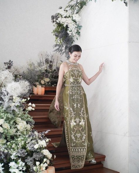 a woman in a long, green, intricately patterned dress stands on a wooden staircase. she is smiling and holding the train of her dress. the staircase is decorated with white flowers and greenery.  the lighting is soft and natural.