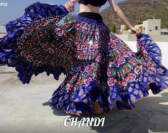 a woman is wearing a vibrant, full, and flared blue skirt with intricate mirror work and colorful patterns, suggestive of traditional rajasthani or indian bohemian style clothing. she is dressed in a black top and standing outdoors on a concrete surface. the skirt is designed for belly dancing or other forms of dance.