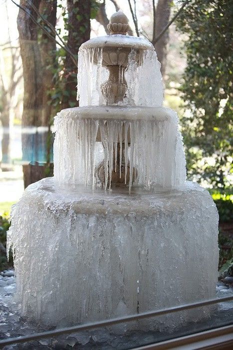a threetiered fountain is completely covered in ice and icicles. the fountain is outdoors, surrounded by trees and greenery, suggesting a garden or park setting. the image depicts a cold, winter scene with the fountain frozen solid.