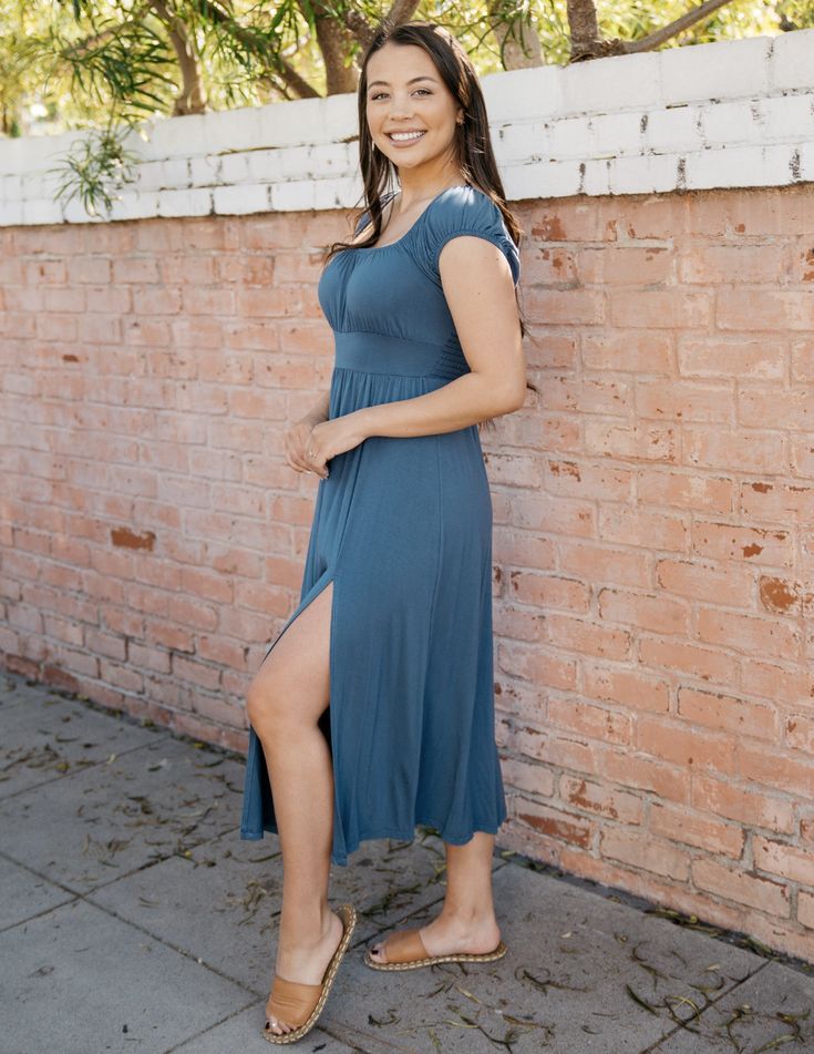 a woman in a blue dress is standing in front of a brick wall. she is wearing brown sandals and has her hair down. the dress has a slit up the leg and short sleeves.  she is smiling and looking over her shoulder.