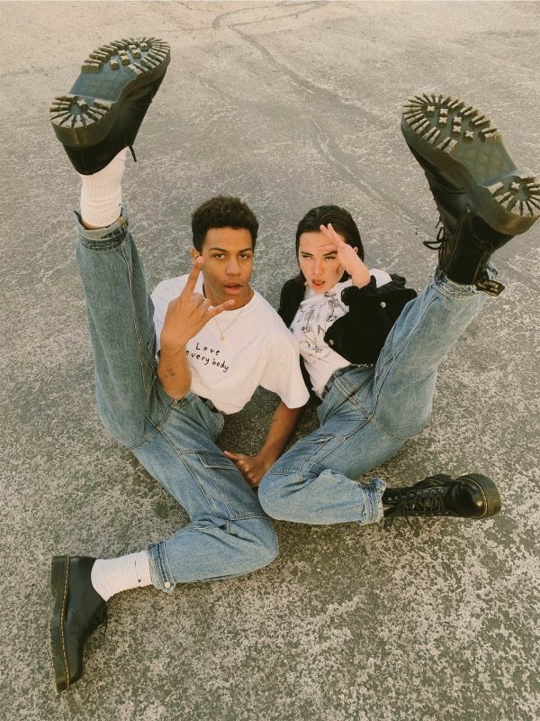 two young adults, a male and a female, are sitting on a concrete surface. the male is wearing a white shirt and blue jeans with black boots. the female is wearing a black jacket, blue jeans, and black boots. the male has his leg up in the air, and the female has her leg up in the air. they are both smiling and looking at the camera.