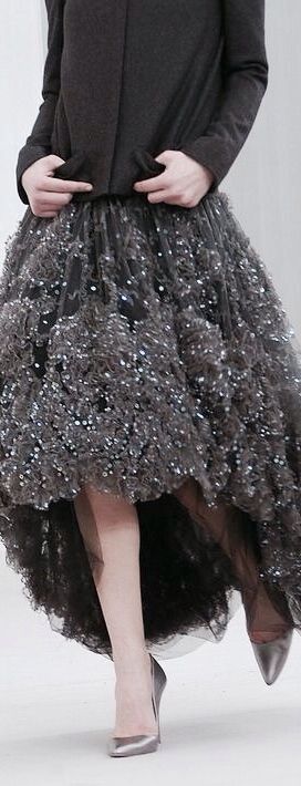 a stylish woman in a black dress and silver high heels poses on a runway. her dress features a highlow hemline with a black bodice and a sequined skirt that cascades down the front. her dress is both elegant and stylish, and her pose adds to the overall glamor of the look.