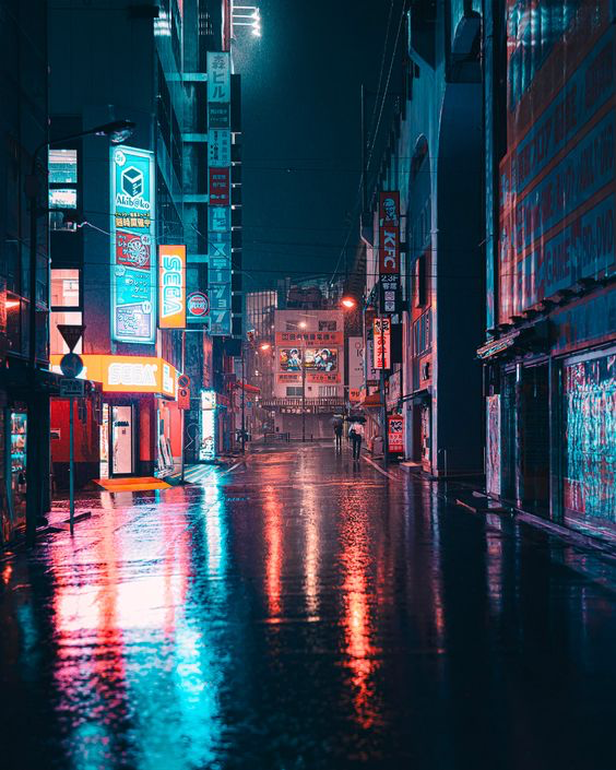 an image of a city street at night. the street is wet from rain, and the neon lights from the buildings are reflected in the puddles. the buildings are tall and narrow, and there are people walking on the sidewalk. the overall atmosphere is dark and moody, with a futuristic, cyberpunk vibe.