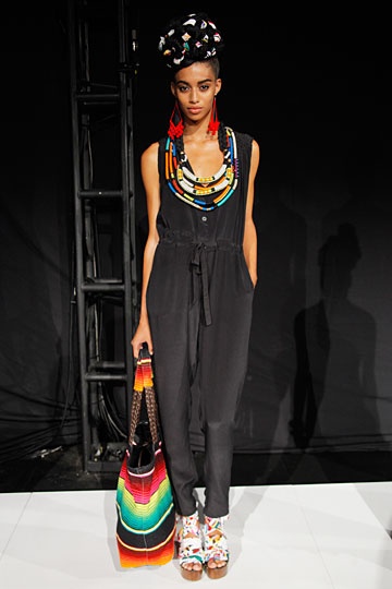 a young black woman wearing a black jumpsuit, a large colorful beaded necklace, and matching sandals walks a runway.  she is holding a colorful bag.  she has a trendy hairdo, with hair pulled back into a bun with colorful objects attached to her hair.
