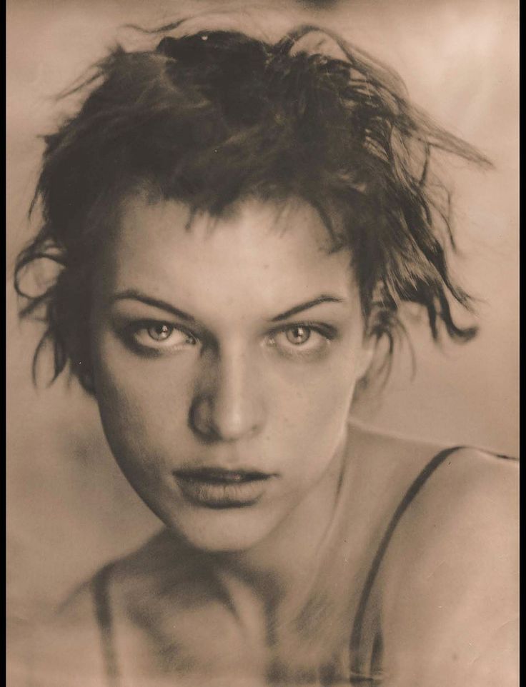 a black and white portrait of milla jovovich. she is looking directly at the camera with a serious expression. her hair is short and messy. she is wearing a simple top with thin straps.