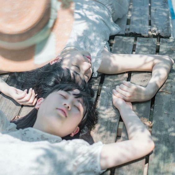 two girls are lying on a wooden plank surface, with their arms intertwined. they appear to be sleeping peacefully, bathed in dappled sunlight.  the image evokes a sense of serenity and summery relaxation.