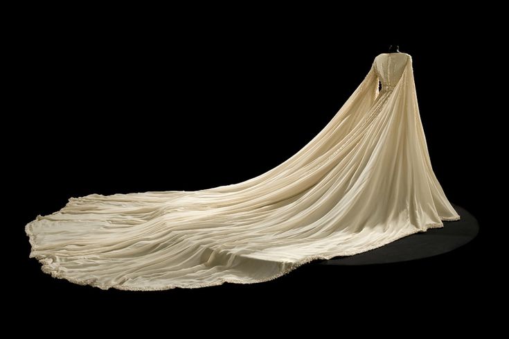 a fulllength shot shows a long, ivorycolored vintage wedding dress on a mannequin against a black background. the dress has long sleeves, a fitted bodice, and a very long train that fans out across the floor. a cape detail is attached at the shoulders and falls behind the dress. the dress and train are intricately pleated. the train has a decorative edge. the dress is welllit, making the details visible.