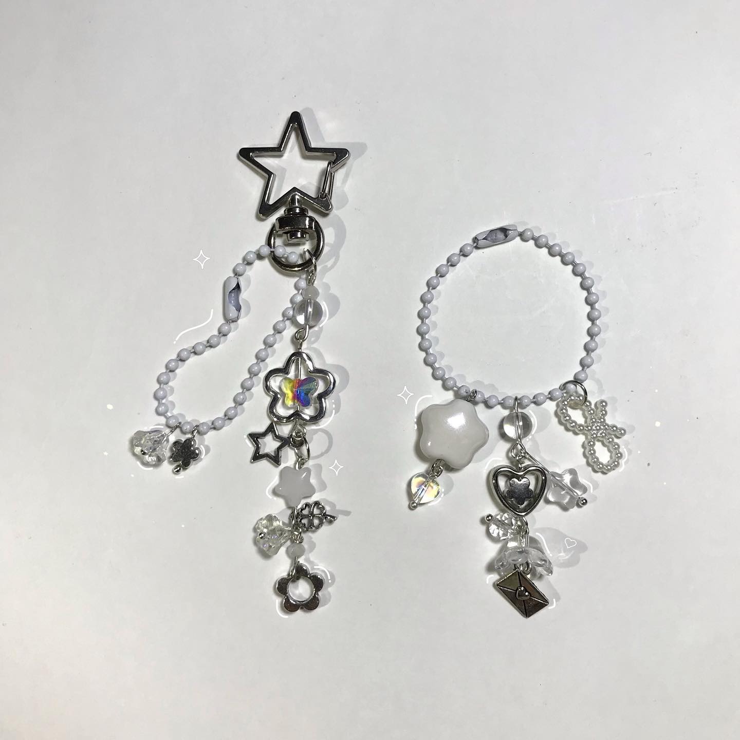 the image contains two handmade keychains with white beads and various charms. one keychain has a large starshaped clasp at the top and hangs a variety of charms, including a flower with iridescent petals, a small star, a flowershaped bead, and a small metal flower. the other keychain is circular, made entirely of white beads, and hangs charms such as a white cloudshaped bead, a heart, an envelope and beads.