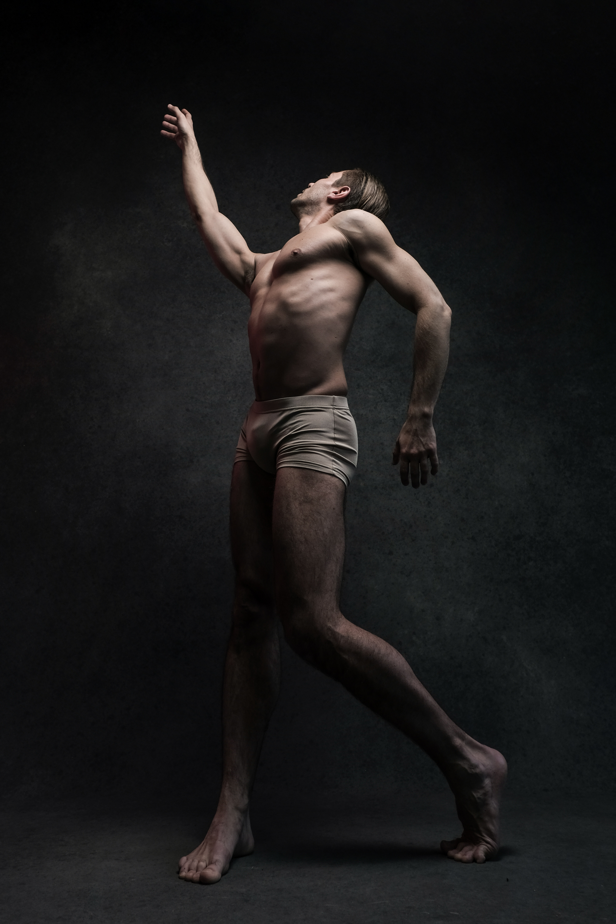 a shirtless male dancer poses dramatically in front of a black background. he reaches his left arm upward as if reaching for something unseen, and his pose emphasizes the length of his body and limbs. the lighting on the dancer is dramatic and highlights his physique.