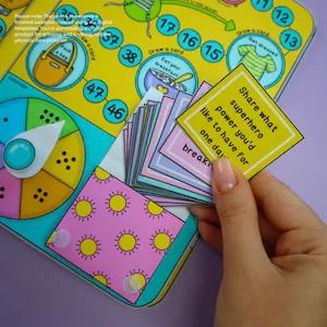 a closeup shot shows a board game with colorful illustrations and numbered spaces. a stack of cards with pastel borders sits on the board, and a hand holds a yellow card with the text "share what superhero power you'd like to have for one day." the game includes a spinner and various action prompts on the board.