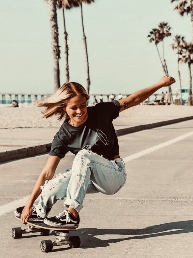 a young blonde woman is skateboarding along a beachside path. she is wearing a dark tshirt, light wash ripped jeans, and black vans shoes. the background features palm trees, a sandy beach, and the ocean in the distance. the overall mood is sunny, carefree, and stylish.