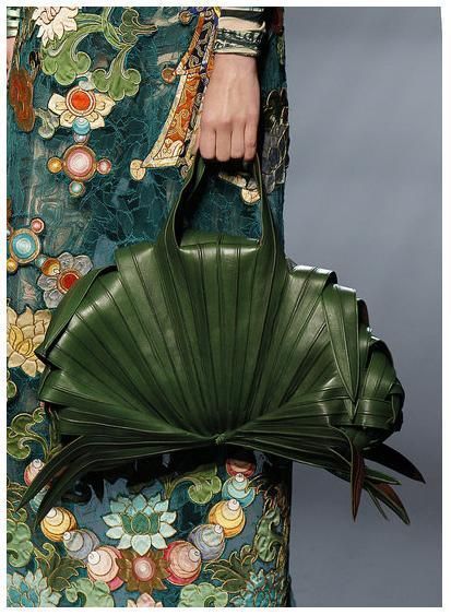 a woman in a teal floral embroidered dress is holding a unique green palm leaf handbag. the bag has a fanlike shape and is made of intricately woven palm leaves. the woman's hand is holding the top of the bag strap. the image is a stylish and elegant fashion moment.