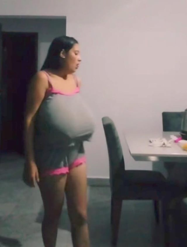 a young woman wearing a pink and gray tank top and pink shorts is standing in her kitchen with her back to the camera looking away. she has long dark hair and is looking away from the camera. in the background, there is a table and chair. the room is well lit and there is a doorway in the background.  the woman looks relaxed and comfortable as she stands in her home.