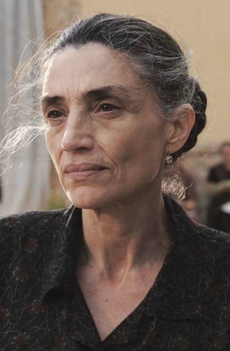 a portrait of an older woman with gray hair pulled back into a bun. she has wrinkles on her face and is wearing a patterned shirt. she has a small earring in her left ear. the background is blurred.