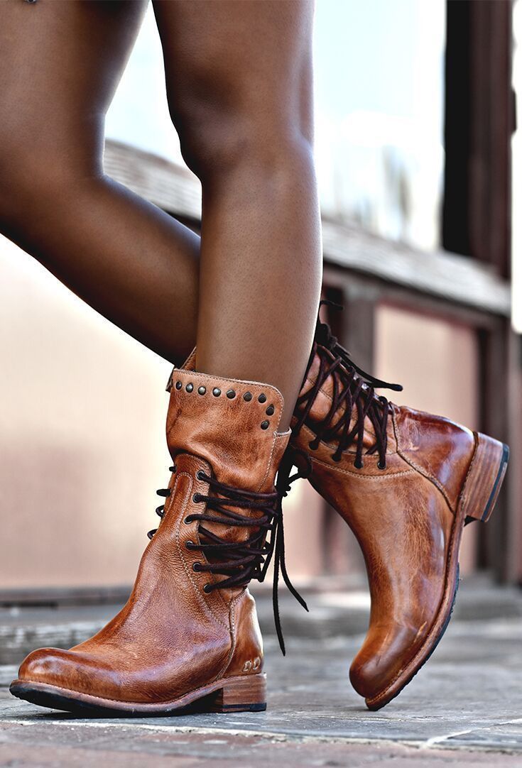closeup shot showcases a woman's legs clad in a pair of stylish brown leather boots. the boots are midcalf length, featuring a laceup front and subtle stud detailing along the topline. the leather appears to be a distressed or vintage style, adding to the boots' rustic charm. the woman's legs are positioned in a way that highlights the boots' silhouette and design. the background is blurred, suggesting an outdoor setting. the overall aesthetic leans toward a bohemian or western style.