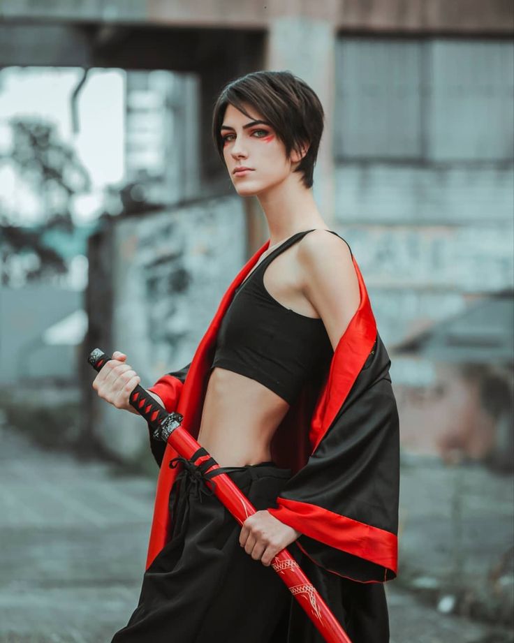 a woman dressed in cosplay attire, holding a red katana sword, stands in an urban outdoor setting. she wears a black crop top, black pants, and a black and red robe. her hair is short and she has red makeup around her eyes.
