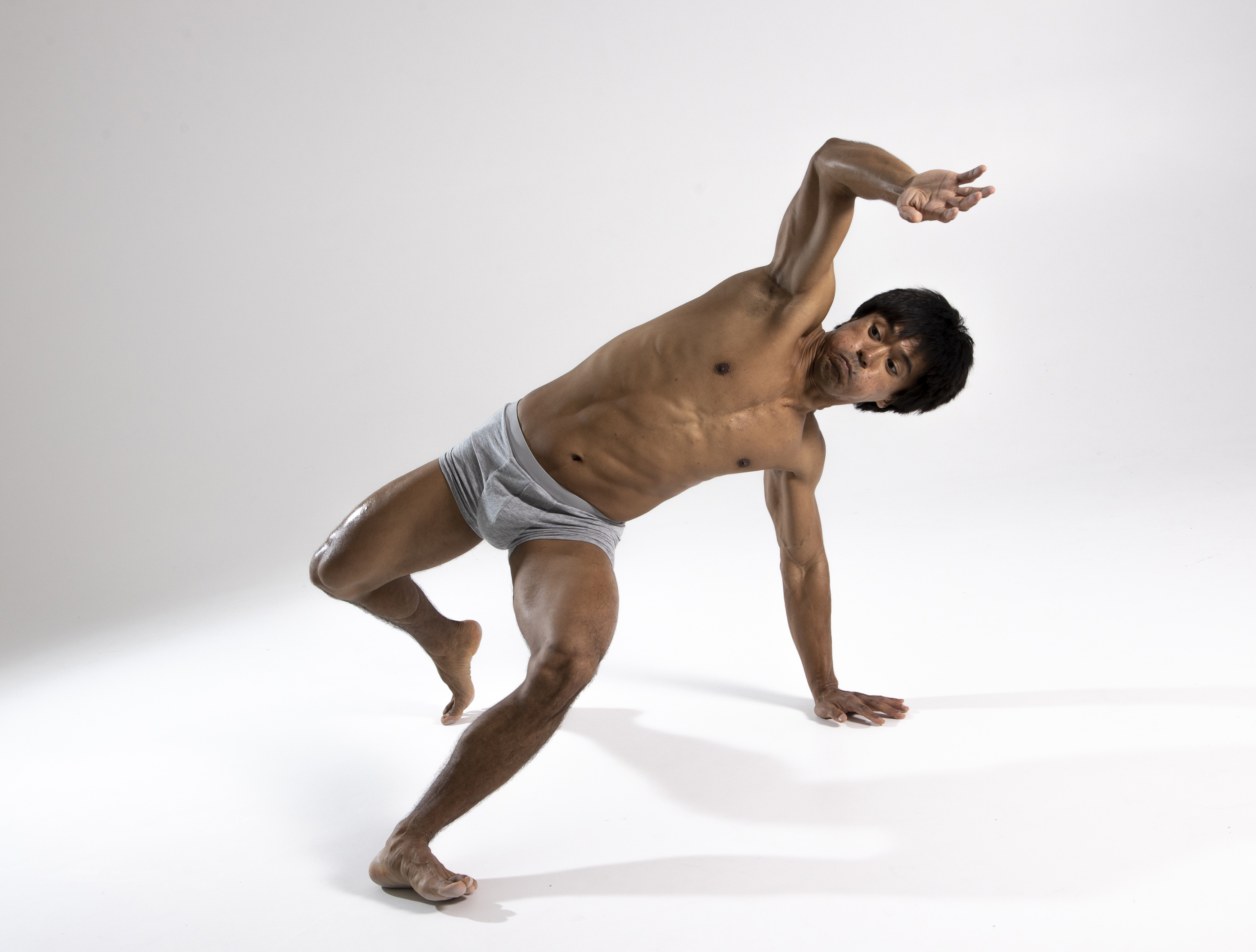 a shirtless man in gray briefs is captured middance move against a plain white background. he's barefoot, leaning to his right, with his right hand touching the floor for support and his left arm extended upwards. the pose showcases his flexibility and athletic build.