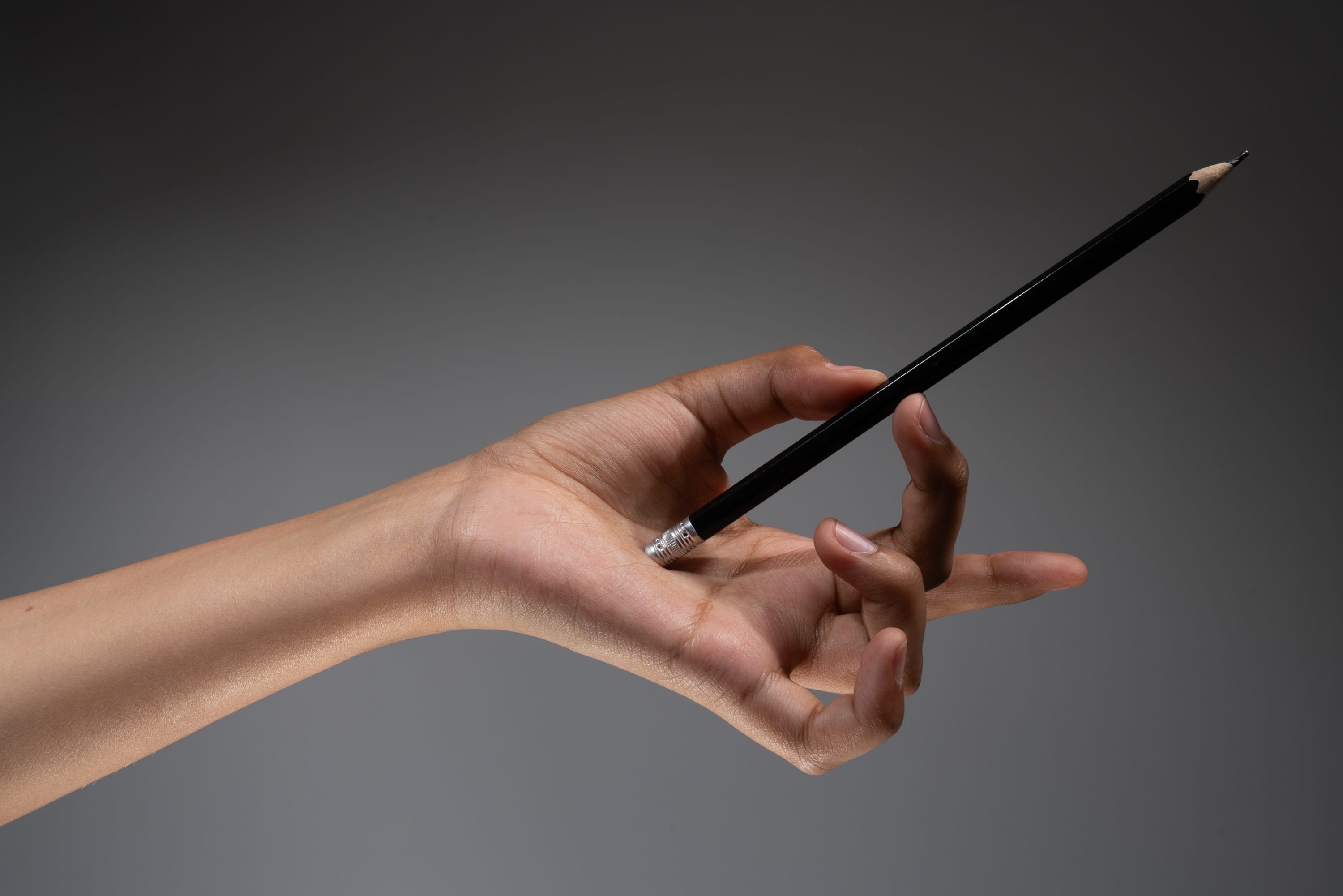 the image shows a hand holding a black pencil against a gray background. the pencil is held between the thumb and index finger, with the point facing upward. the hand is slender and has a light brown complexion. the gray background is smooth and even. the lighting is soft and diffused, creating a subtle shadow on the hand.
