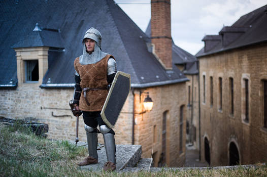 a medieval knight stands in a stone village street, holding a shield and sword. he is wearing chainmail, a leather tunic, and a helmet. the background shows old stone buildings. the scene evokes a historical or fantasy setting. the image is a professional photograph, welllit and sharply focused.