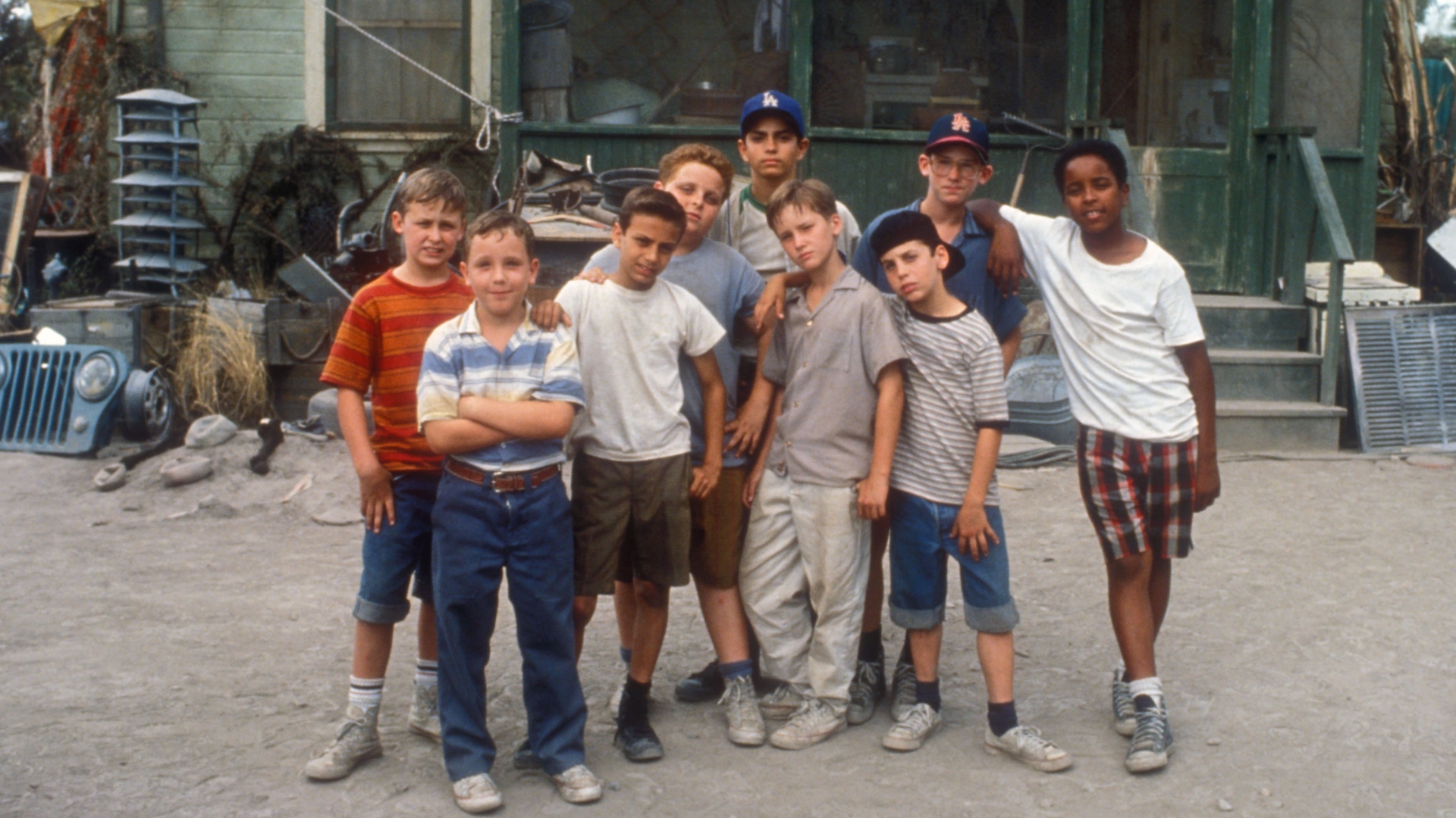 a group of eight boys stand in a line in front of a house. they are all wearing casual clothes and have different hairstyles. the boy in the center is wearing a white shirt and blue jeans. the boy on the left is wearing a striped shirt and blue shorts. the boy on the right is wearing a white shirt and plaid shorts. the house is old and has a porch. there is a dirt road in front of the house. the photo is taken from an eye level perspective and the lighting is natural and sunny. the photo evokes a sense of friendship, childhood, and summer.
