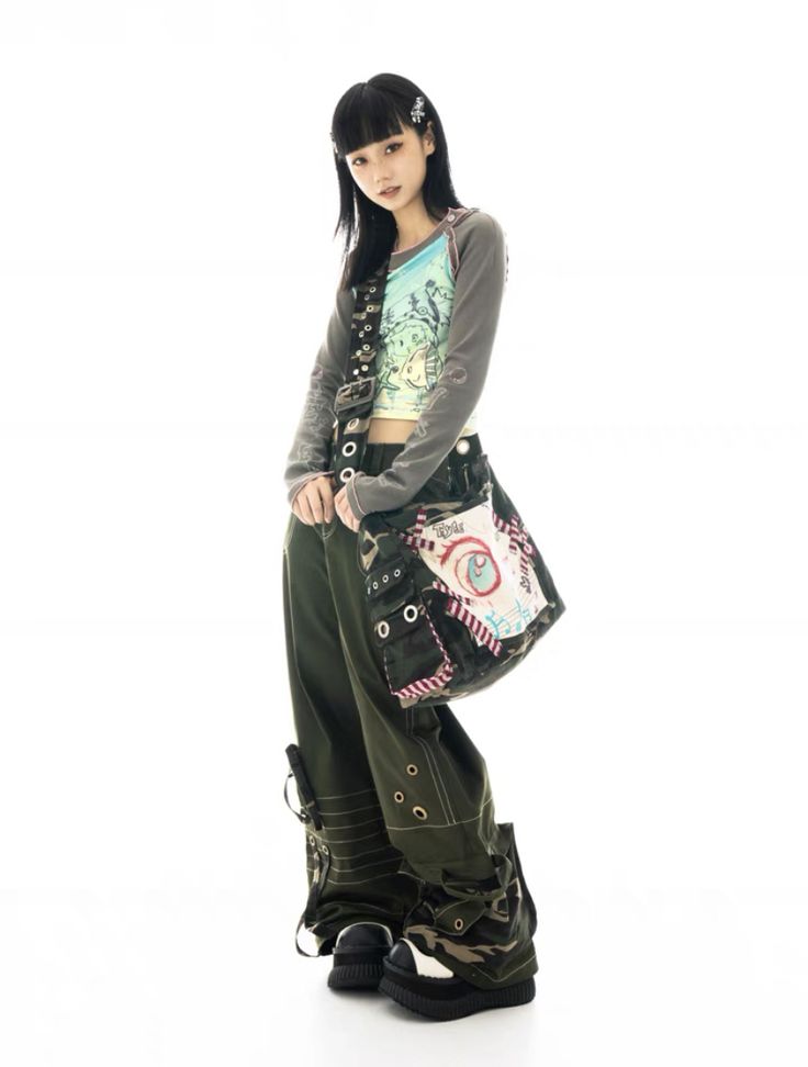 full shot of a young woman with dark hair and bangs posing in a studio against a white backdrop. she's wearing a longsleeved graphic tee, olive green cargo pants with camouflage detailing at the bottom, and platform boots in a similar camouflage pattern.  a distinctive patchwork bag with an eyelike design hangs from her shoulder. the overall style is a blend of streetwear, grunge, and alternative fashion.  her pose is relaxed but styled.