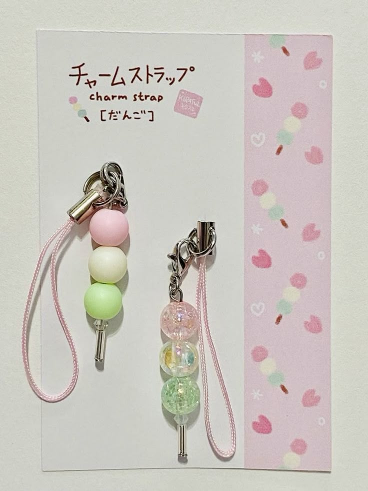 the image shows two dango charm straps displayed on a white card with a pink border. the card features the text "charm strap" and "[dango]" in japanese, along with a small illustration. the charm straps consist of three colorful beads arranged to resemble a dango skewer. one charm strap has solid pastel beads in pink, white, and green. the other charm strap features crackled transparent beads in pink, clear, and green. both charm straps have silver hardware and pink straps. the pink border on the card is decorated with dango and cherry blossom illustrations.