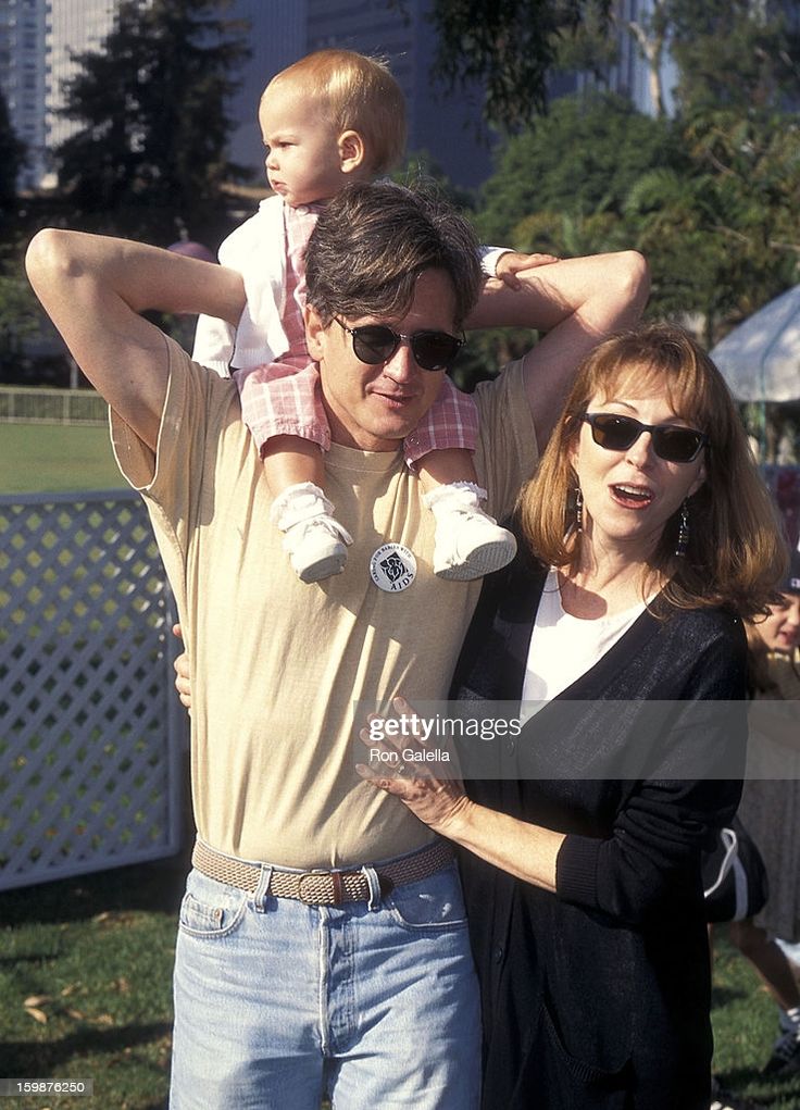 a photo of a family at a park with the father carrying the young daughter on his back. the mother is standing next to her husband and daughter with her arms around them.