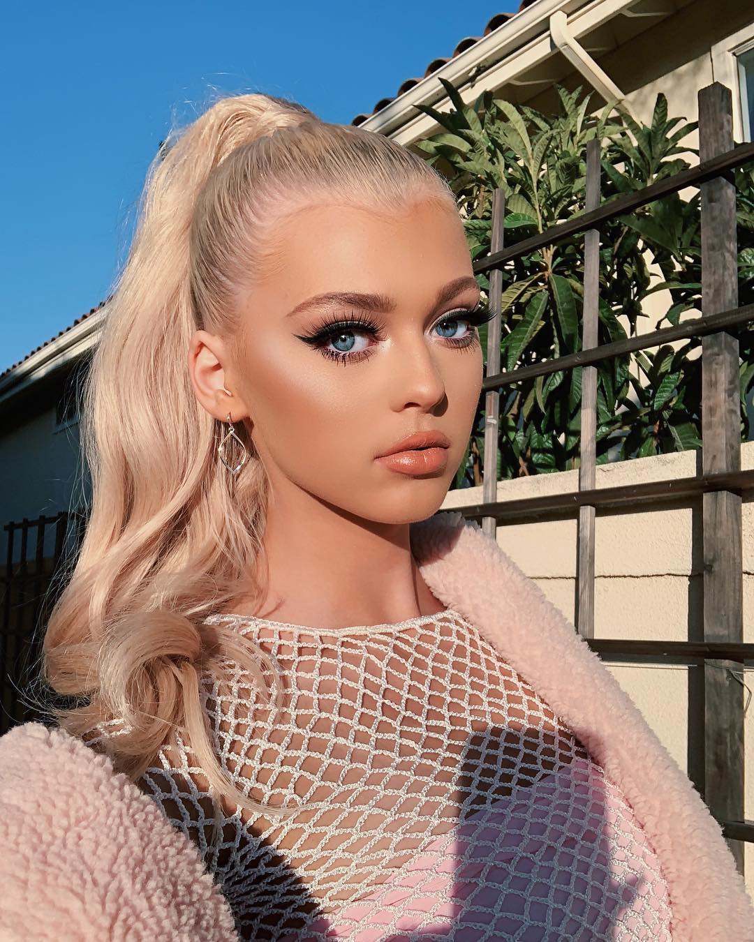 a young woman with blonde hair in a high ponytail and wearing heavy makeup, including winged eyeliner and glossy lips, is posing outdoors. she's wearing a white mesh top over a pink shirt and a fluffy pink jacket. she also has diamondshaped earrings. the background includes a fence, lush green plants, a house, and a clear blue sky with bright sunlight.