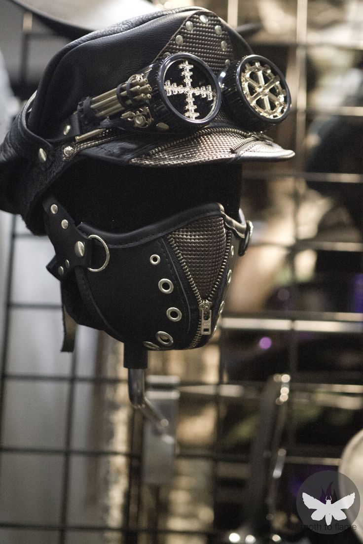 a steampunk mask and cap on a black metal rack. the mask is black leather with silver buckles and a silver zipper. the goggles are attached to the cap, which is black leather with silver accents. the background is blurry, but it appears to be a shop or market.