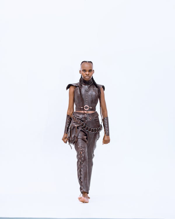 a woman is walking towards the camera, wearing a brown leather outfit consisting of a corset top, highwaisted pants with laceup details, and a chain belt with a small bag attached. she has braids in her hair and is barefoot. she is in a studio with a bright white background.