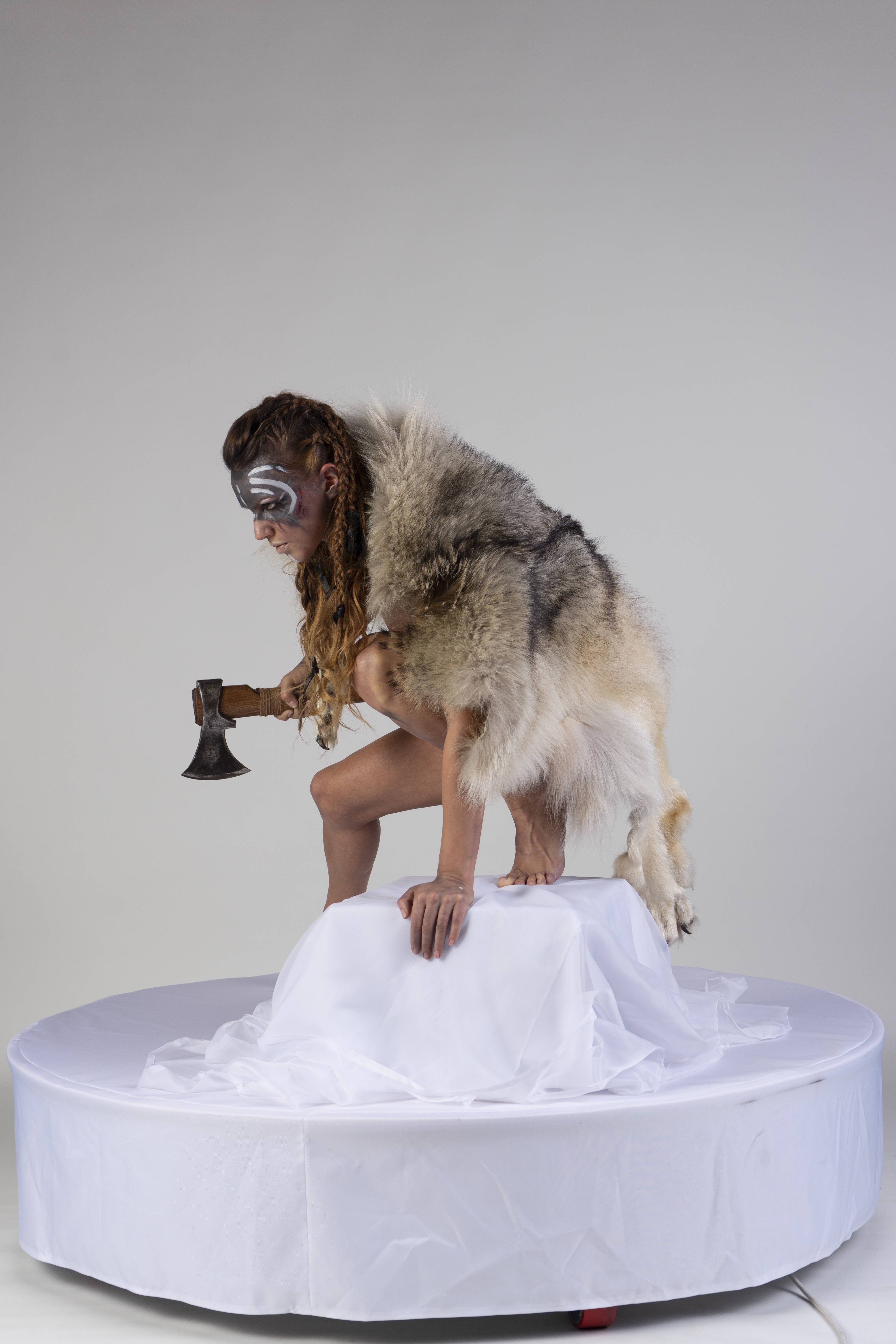 a warrior woman crouches on a stage, holding an axe and wearing a wolf skin. she has body paint on her face. the setting is an indoor studio.