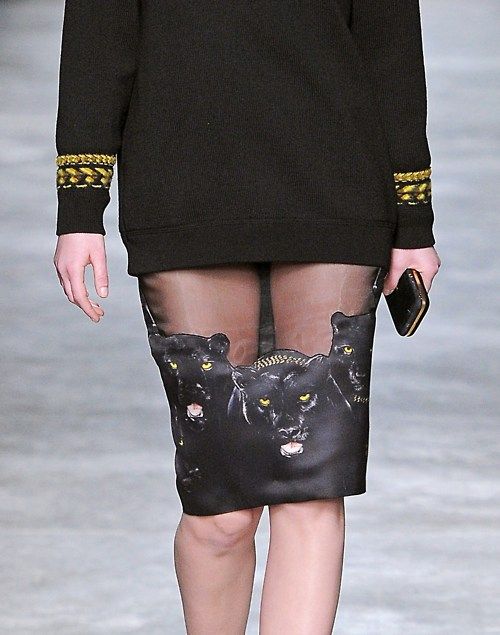 a model on a runway wears a black sweater with gold trim on the sleeves and a skirt with a black panther print on sheer fabric. she is holding a phone in her right hand.