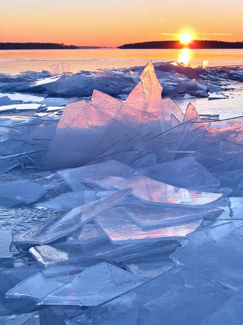 a frozen lake landscape with ice floes at sunset. the image features a closeup of large, transparent ice fragments in the foreground, reflecting the warm hues of the setting sun. in the background, the horizon is marked by a body of water and a distant shoreline, silhouetted against the colorful sky. the overall scene conveys a sense of cold, natural beauty.