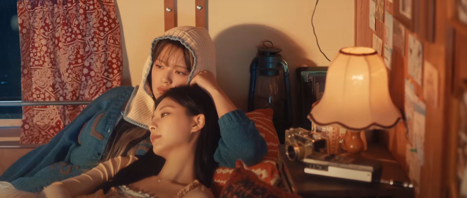 two young women are lying down together in a warmly lit bedroom. one woman is resting her head on the other's shoulder, and they both have serene expressions. the room has a cozy and intimate atmosphere, with a lamp providing soft lighting. the color palette is warm and inviting.